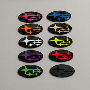 Könnte beinhalten: Zehn ovale Embleme mit schwarzem Hintergrund, jeweils mit einem Stern-Design in verschiedenen Farben: Pink, Blau, Orange, Gelb, Weiß, Lila, Rot, Grün und Schwarz. Wahrscheinlich Auto-Embleme oder Zubehör.