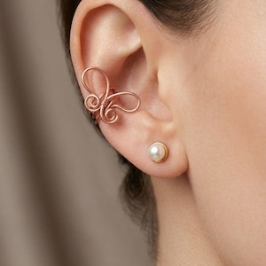May include: A rose gold-colored wire ear cuff shaped like a butterfly with swirling wire details, paired with a single round white pearl stud earring set in a gold-colored metal base, both worn on the earlobe and cartilage area.