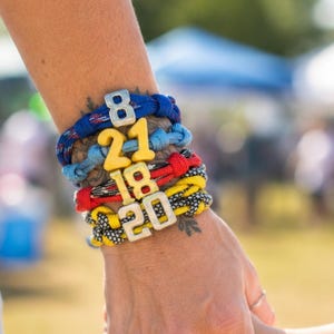 May include: A stack of four woven cord bracelets in blue, red, and yellow, each featuring a prominent number charm. The charms display the numbers 8, 21, 18, and 20 in a mix of metallic silver and solid yellow plastic finishes.