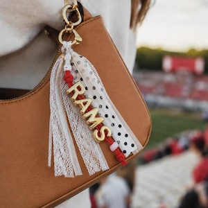May include: A bag charm featuring a gold-toned metal clasp, a red cord, and gold-toned letters spelling RAMS. The charm includes white lace strips and a white ribbon with black polka dots, attached to a tan leather-like handbag with a gold zipper.