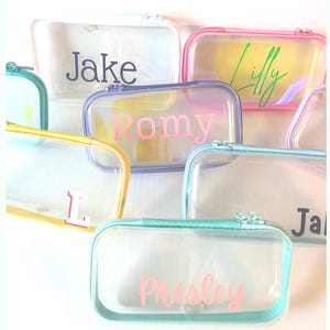 Könnte beinhalten: Eine Sammlung rechteckiger, durchsichtiger Kunststoffetuis mit farbigen Reißverschlüssen und Rändern. Die Etuis sind mit personalisierten Namen oder Initialen wie Jake, Lilly, Romy, L und Presley in verschiedenen Schriftarten und Farben bedruckt.