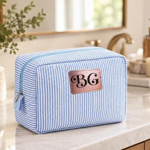 May include: A rectangular toiletry bag featuring a light blue and white pinstripe pattern. The bag has a light blue zipper and a centered, rectangular rose gold metallic patch embossed with the black capital letters BG in a script font.