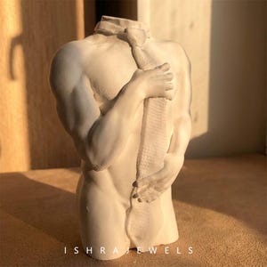 May include: A white matte sculpture of a human torso featuring a necktie draped from the collar area down the centre of the chest. The figure has defined musculature and arms positioned across the torso. The text ISHRAJEWELS is at the bottom.