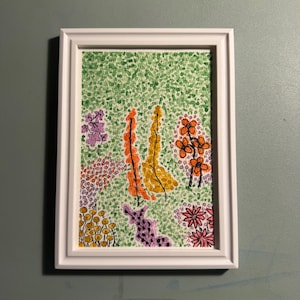 Op de afbeelding: Een ingelijste aquarel van een tuin. Het kunstwerk toont kleurrijke bloemen en bladeren in tinten oranje, geel, paars en roze tegen een groene, gestippelde achtergrond. De schildering zit in een witte lijst.