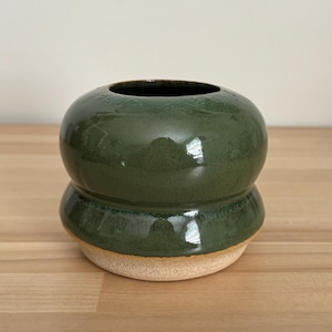 Puede incluir: Un jarrón de cerámica verde oscuro hecho a mano con un diseño apilado único. El jarrón tiene un acabado brillante y se asienta sobre una base natural sin esmaltar. La abertura superior es oscura, contrastando con el cuerpo verde.