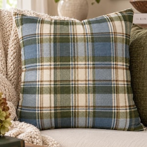 Pode incluir: Uma almofada quadrada com um padrão xadrez em tons de verde sálvia, azul ardósia, creme e castanho. O tecido tem uma textura visível e o design consiste em linhas horizontais e verticais que se cruzam formando uma grelha.