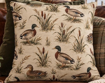 Op de afbeelding: Een vierkant sierkussen met een beige stoffen hoes met een herhalend patroon van wilde eenden, lisdodden en hoog gras in bruin-, groen- en tinten. Het kussen heeft een zichtbare geweven textuur en is stevig gevuld tot een volle vorm.