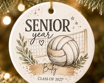 May include: A round white ceramic ornament featuring a volleyball graphic, a net, and green leaf accents. The text reads SENIOR year, Bailey, and CLASS OF 2027, with a small heart icon. It hangs from a gold string against a blurred evergreen background.