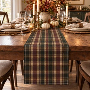 Puede incluir: Un camino de mesa rectangular con un patrón de cuadros en tonos verde oscuro, burdeos y beige. El camino de tela está colocado sobre una mesa de comedor de madera, entre cubiertos con servilletas blancas y cristalería.