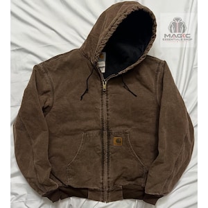 May include: A brown hooded jacket with a full-length front zip, a black interior lining, and ribbed cuffs. The garment features a front pouch pocket with a small square Carhartt brand patch. A logo for Magic Essentials Shop is visible.