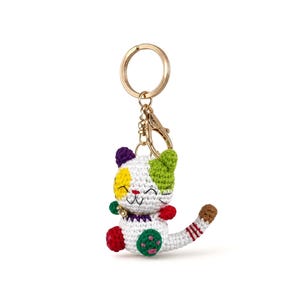 May include: A crocheted cat keychain featuring a white body with multicolored patches in yellow, green, purple, and red. The cat has a stitched smiling face, a small bell around its neck, and a long tail with red stripes, attached to a gold metal ring.
