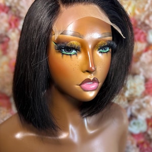 May include: A dark brown bob wig with a lace front is displayed on a mannequin. The mannequin has dramatic makeup with gold eyeshadow and green eyeliner. The background is a soft focus of pink and white flowers.