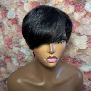 May include: A black wig with a short, layered style and a side-swept bang. The wig is styled on a mannequin head with a painted face, including pink lips and freckles. The background features a wall of pink and white flowers.