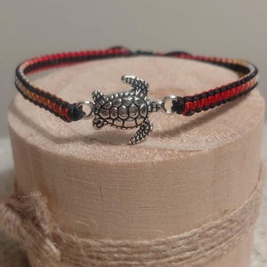 Könnte beinhalten: Ein handgefertigtes Armband mit einem silbernen Schildkrötenanhänger. Das Armbandband ist schwarz, rot und beige. Der Schildkrötenanhänger ist mit silbernen Verschlüssen am Band befestigt. Das Armband wird auf einem Holzblock präsentiert.