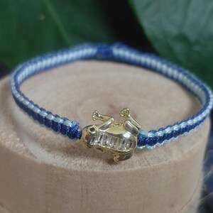 May include: A bracelet featuring a gold-toned frog charm with clear baguette-cut stones set into its back. The band is constructed from braided cord in alternating shades of dark navy blue and light sky blue, creating a woven pattern.