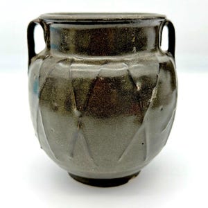 May include: A dark brown ceramic vessel with a rounded body, a wide neck, and two vertical handles on opposite sides. The exterior surface features a glossy glaze with a subtle, textured pattern of diagonal indentations around the midsection.