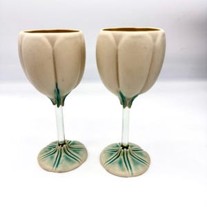 Puede incluir: Dos copas de cerámica con cuenco y base en forma de tulipán de color crema mate, acentuadas con detalles en verde azulado en la unión del tallo y la base. Cada copa tiene un tallo de vidrio transparente que une el cuenco con la base.