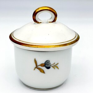 May include: A white porcelain sugar bowl with a matching lid featuring a gold-trimmed handle and rim. The body of the bowl is decorated with a small, centred floral motif consisting of a dark grey flower with gold-painted leaves and stems.