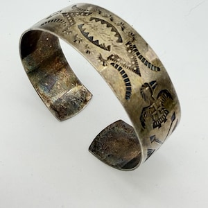 May include: A silver cuff bracelet featuring a wide band with a tarnished finish and intricate stamped designs. The surface displays detailed engravings of a bird figure, geometric patterns, and linear motifs across the exterior metal surface.
