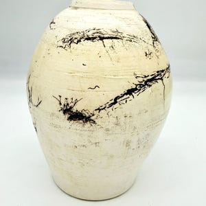 May include: A cream-colored ceramic vase with a wide, rounded body and a narrow neck. The vase features dark, abstract markings across its surface, creating a textured appearance. The vase is a decorative item.