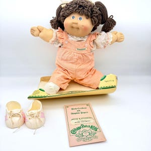 Puede incluir: Muñeca Cabbage Patch Kids con cabello de hilo marrón, ojos azules y mejillas rosadas, vestida con un mono de pana melocotón. Incluye certificado de nacimiento, papeles de adopción y un par de zapatillas blancas con rayas rosas. La muñeca está sentada.