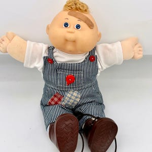May include: A doll with a round face, blue eyes, and curly blonde hair, wearing a white shirt under blue and white pinstriped overalls. The overalls feature red buttons, a red and white plaid patch, and a blue and white plaid patch. Brown shoes.