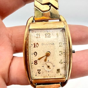 May include: A vintage gold-tone Bulova wristwatch with a rectangular case and a matching gold-tone link bracelet. The off-white dial features gold-tone Arabic numerals, gold-tone hands, and a small sub-dial for seconds at the six o'clock position.