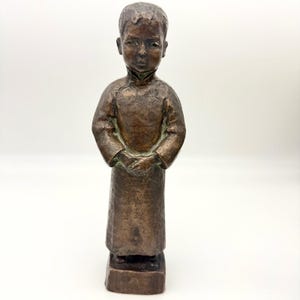 May include: A bronze sculpture of a young child in a long-sleeved garment, hands clasped in front. The statue has a textured surface and stands on a rectangular base. The figure's expression is solemn.