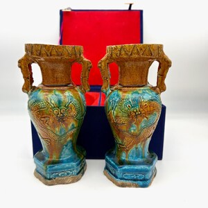 Puede incluir: Un par de jarrones antiguos de cerámica con un motivo de dragón. Los jarrones presentan una parte superior marrón, asas y detalles de dragón, con un esmalte turquesa y marrón. Se exhiben frente a una caja azul con interior rojo.