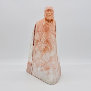 May include: A carved stone sculpture of a figure wearing a head covering and draped garment. The sculpture is primarily peach and white, with a carved butterfly and floral design on the front. The figure has a serene expression.