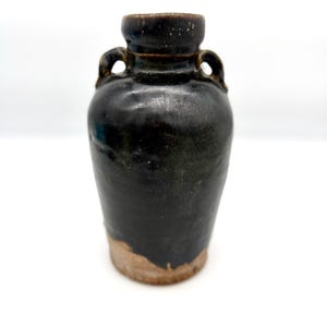 May include: A dark brown, glazed ceramic vase with a rounded body, a narrow neck, and two small handles on the sides. The base of the vase is unglazed, revealing a light tan, textured clay surface that contrasts with the glossy dark finish above.