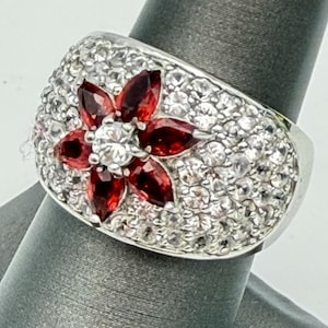May include: A wide silver-toned ring featuring a central flower motif composed of six marquise-cut deep red stones surrounding a single round clear stone. The entire surface of the band is densely encrusted with numerous small clear stones.