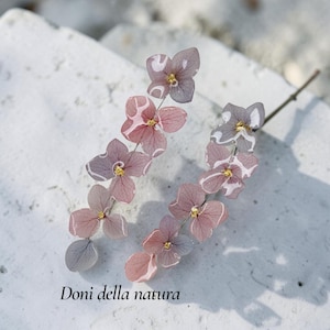 Peut inclure: Une paire de boucles d'oreilles composées de pétales d'hortensia pressés dans des tons rose poudré et gris doux, ornés de petits centres dorés. Les pétales présentent une finition blanche marbrée translucide. Le texte Doni della natura est visible.