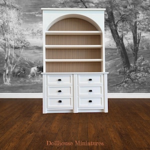 May include: A white dollhouse miniature hutch with a curved top and three shelves. The back of the hutch has vertical wooden slats. The bottom section has six drawers with black knobs. The text "Dollhouse Miniatures" is at the bottom.