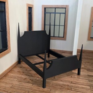 May include: A black dollhouse bed frame with a tall headboard and footboard. The bed frame is made of wood and has a simple, elegant design. The bed is in a room with wooden floors and windows.
