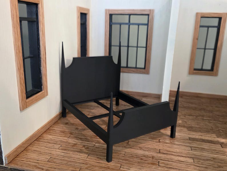 May include: A black dollhouse bed frame with a tall headboard and footboard. The bed frame is made of wood and has a simple, elegant design. The bed is in a room with wooden floors and windows.