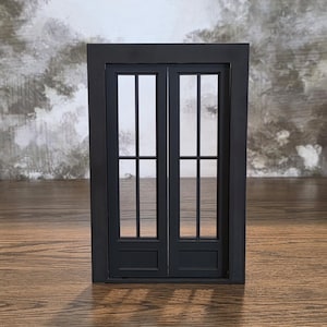 May include: A rectangular black frame containing a pair of double doors. Each door features two vertical glass panes separated by a horizontal muntin, with a solid rectangular panel at the bottom of each door section within the frame structure.