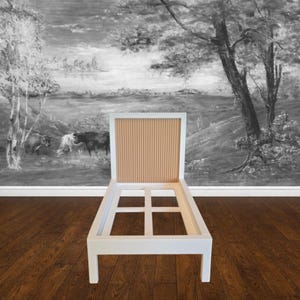 May include: A white wooden chair frame with a tan, vertically-lined back. The chair is set against a black and white mural of a pastoral scene with trees and grazing animals. The chair sits on a dark wood floor.