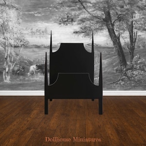 May include: A black dollhouse bed frame with a tall, pointed headboard and footboard. The bed frame is made of wood and has a minimalist design. The background features a black and white landscape painting. The bed frame is a miniature furniture item.