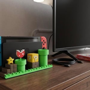 May include: A Nintendo Switch console dock featuring a Super Mario theme with a green pipe, a yellow question mark block, a brown brick staircase, a yellow star, and a red and white piranha plant, all set on a green grass-textured base.