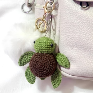 Puede incluir: Un llavero de tortuga hecho a mano con ganchillo en tonos verdes y marrones, sujeto a un cierre dorado. La tortuga tiene un cuerpo redondo y extremidades en forma de aleta. El llavero está sujeto a un bolso de color claro.