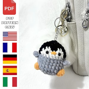 May include: A crochet penguin keychain with a black head, white face, gray body, and orange feet, attached to a silver metal ring. The image includes a red PDF icon, the text PDF PATTERN ONLY, and flags for the United States, France, Germany, Spain, and Italy.
