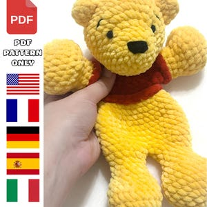 May include: A yellow and red crocheted bear plush toy with a textured yarn finish, black eyes, and a black nose. The image includes a red PDF icon, the text "PDF PATTERN ONLY," and a vertical column of flags representing the United States, France, Germany, Spain, and Italy.