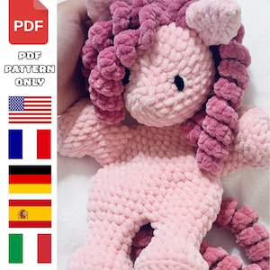 Puede incluir: Un unicornio de ganchillo rosa claro hecho a mano con pelo rizado y cola rosa oscuro. El unicornio tiene ojos de botón negros y un aspecto suave y texturizado. La imagen incluye un icono PDF y el texto "PDF PATTERN ONLY".