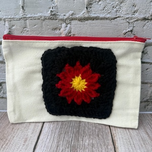 May include: A rectangular off-white canvas pouch featuring a red zipper closure. Centered on the front is a square black crochet patch with a vibrant red and yellow flower motif stitched into the middle of the dark yarn square.