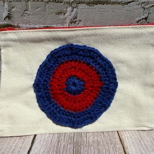 May include: A rectangular off-white canvas zipper pouch featuring a circular crochet appliqué centered on the front. The crochet design consists of a dark blue center, a vibrant red middle ring, and a thick dark blue outer border.