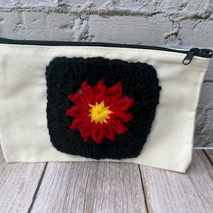 May include: A rectangular off-white canvas zippered pouch featuring a square black crochet patch centered on the front. The crochet patch displays a vibrant red flower design with a bright yellow center, adding a textured contrast to the fabric.