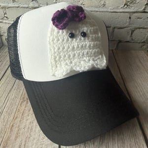 May include: A white and black trucker hat featuring a crocheted white ghost applique on the front panel. The ghost has two small black bead eyes and a dark purple crocheted bow attached to the top of its head, centered on the white foam crown.