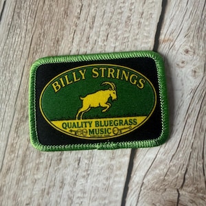 May include: A rectangular embroidered patch with a green border and black background. It features a central green oval containing a yellow goat illustration, the text BILLY STRINGS, and the text QUALITY BLUEGRASS MUSIC with small banjo icons.