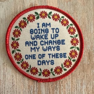 May include: A circular embroidered patch with a red stitched border. The center is off-white with blue text that reads: I AM GOING TO WAKE UP AND CHANGE MY WAYS ONE OF THESE DAYS. A wreath of small red flowers with yellow centers and green leaves.